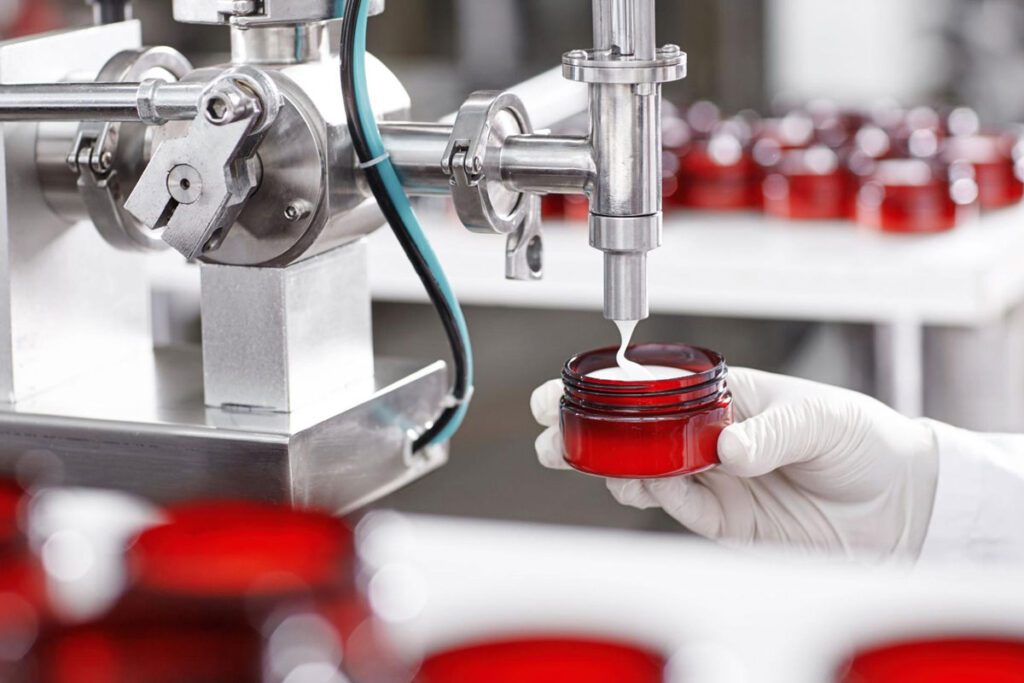Precision dispensing nozzle filling cosmetic cream into a small glass jar on an automated line.